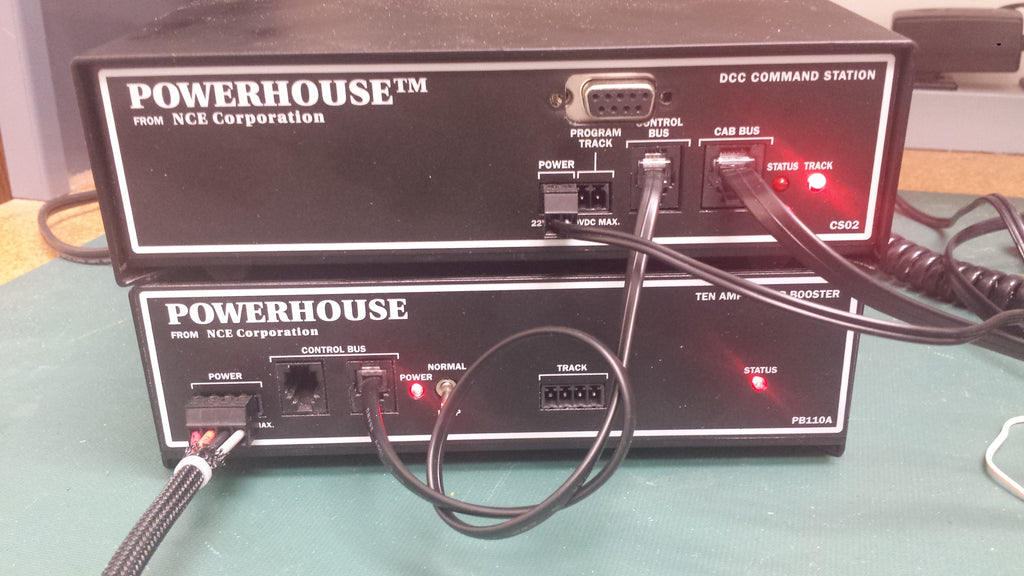 Two black NCE Corporation POWERHOUSE DCC command station units stacked on a green surface, with attached cables and red indicator lights illuminated.