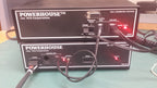 Two black NCE Corporation POWERHOUSE DCC command station units stacked on a green surface, with attached cables and red indicator lights illuminated.